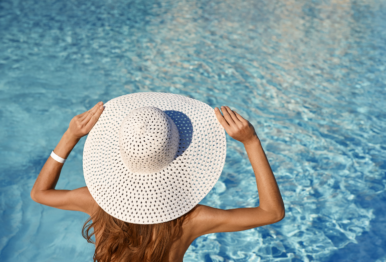 Woman by pool with wide-brimmed hat, using tips from Desert Essence for hair care before and after swimming.