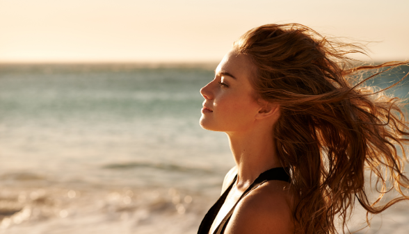 Woman on beach with wind in her hair, epitomizing the perfect summer hairstyle with beachy waves.