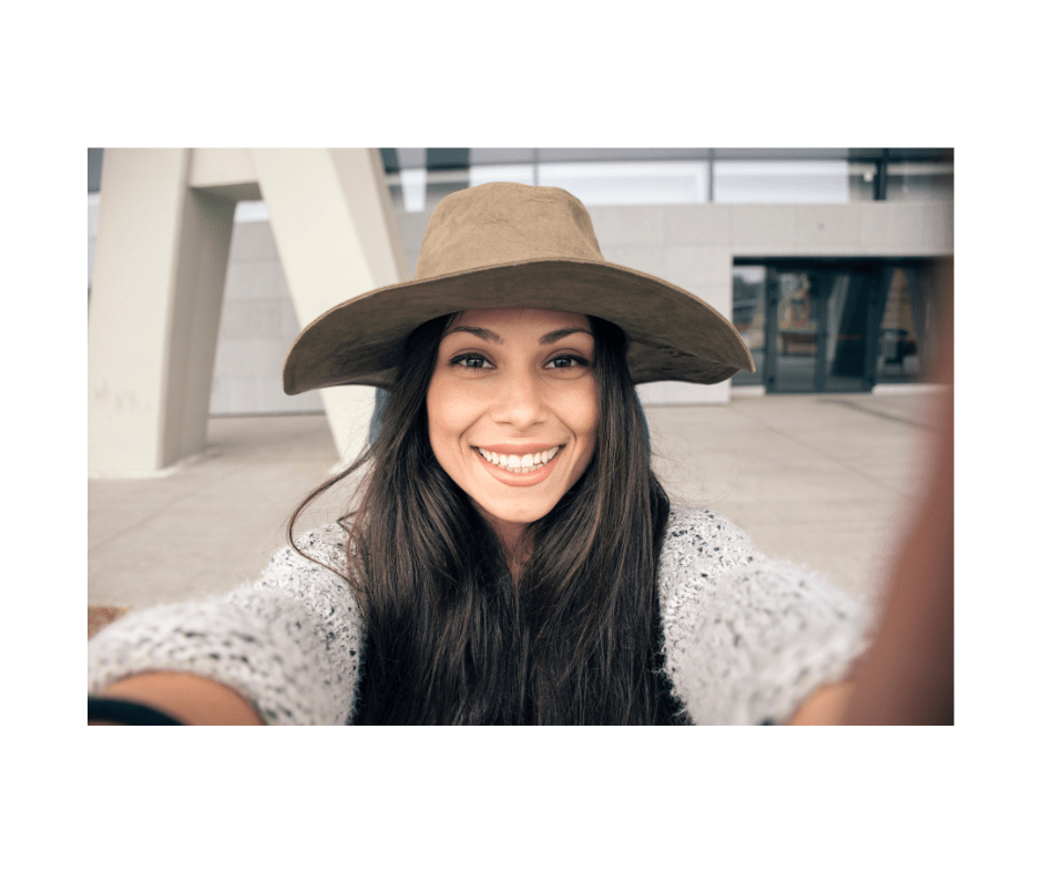 Cheerful woman in a wide-brimmed hat ready for a selfie, radiating flawless skin and hair.