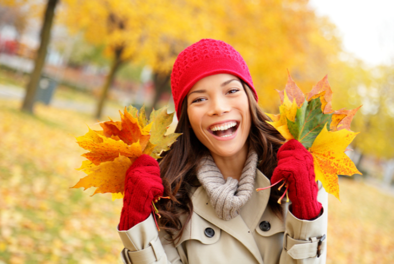 Joyful woman in a red beanie & gloves holding autumn leaves, embodying the spirit of fall, perfect for Desert Essence product highlights.